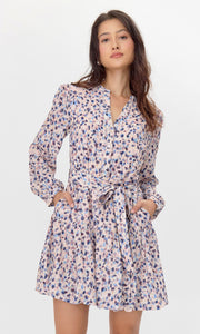 Model wearing a flowy long-sleeve dress with a pink, blue, and purple abstract print. The dress features a tiered skirt, buttoned neckline, and a matching tie belt at the waist. She poses with both hands in her pockets.