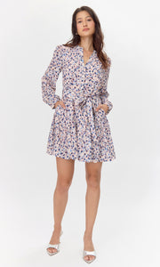 Model wearing a flowy long-sleeve dress with a pink, blue, and purple abstract print. The dress features a tiered skirt, buttoned neckline, and a matching tie belt at the waist. She poses with both hands in her pockets.