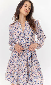 Model wearing a long-sleeve dress with a pink, blue, and purple abstract print. The dress features a tiered skirt, buttoned neckline, and a matching tie belt, which she is adjusting with her hands. She poses with a relaxed expression, looking slightly downward, against a plain white background.
