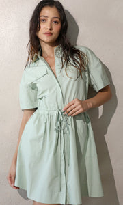 Lifestyle image of model wearing short-sleeve, shirt dress. Leaning against the wall, she shows off the drawstring detail on the waist.